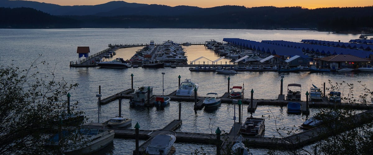A marina with boats docked at piers, surrounded by mountains and a sunset sky.
