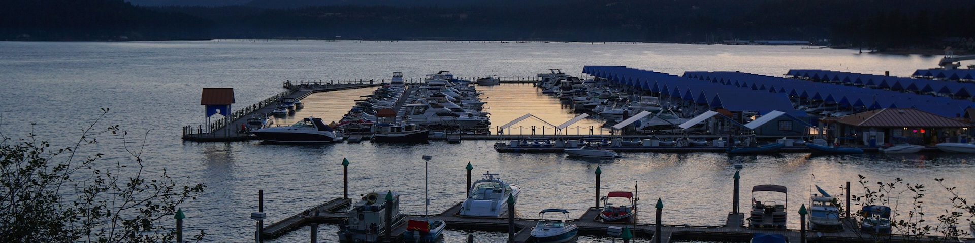 A marina with boats docked at piers, surrounded by mountains and a sunset sky.