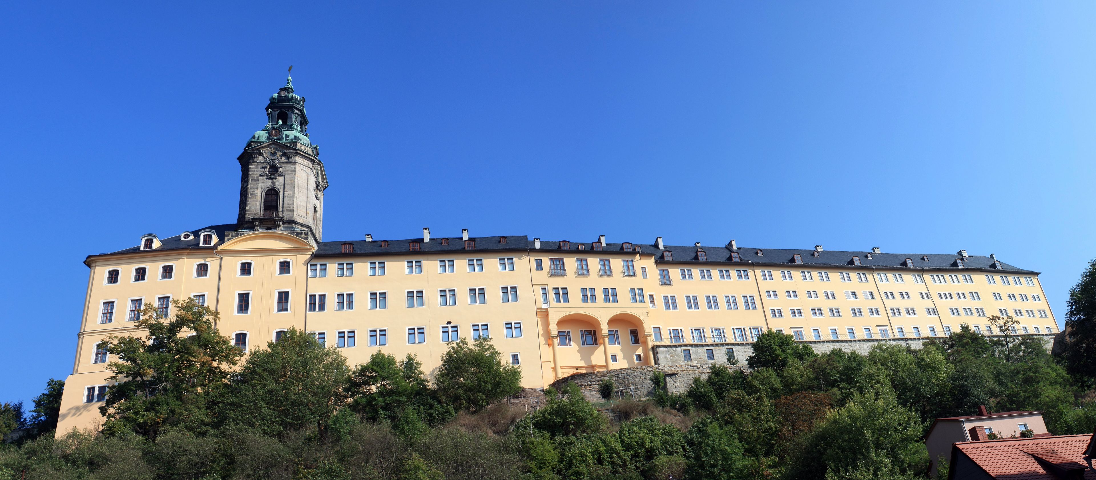 Ein großes, gelbes Gebäude mit einem Glockenturm, umgeben von Bäumen und blauem Himmel.