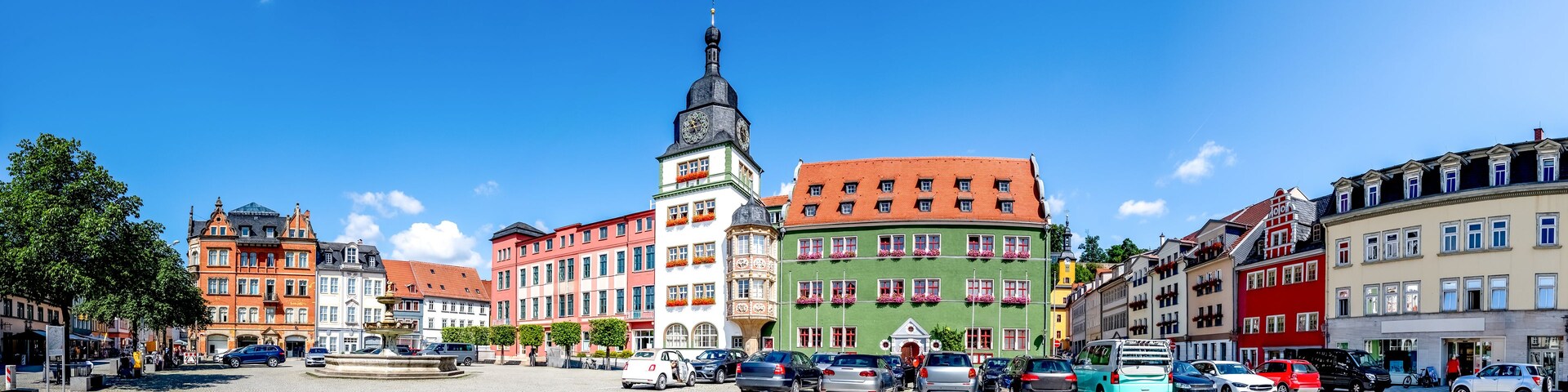 A cobblestone square with colorful historic buildings, a prominent clock tower, and a fountain.