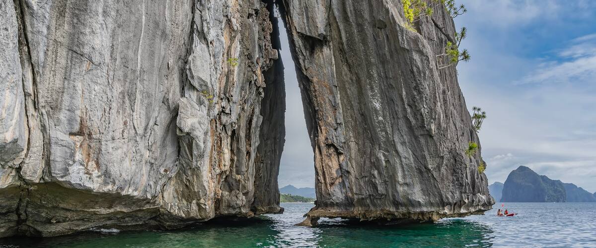 The rock rises above the ocean. There is scant green vegetation on the steep slopes. The narrow gap between the cliffs. A tourist canoe floats nearby. Mountains in the distance. Blue sky, clouds.