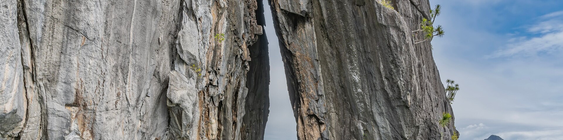 The rock rises above the ocean. There is scant green vegetation on the steep slopes. The narrow gap between the cliffs. A tourist canoe floats nearby. Mountains in the distance. Blue sky, clouds.