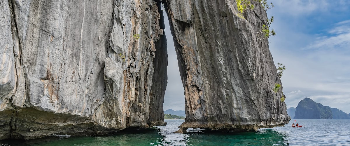 A large rock formation with a narrow passage between two cliffs, surrounded by turquoise water and distant mountains.