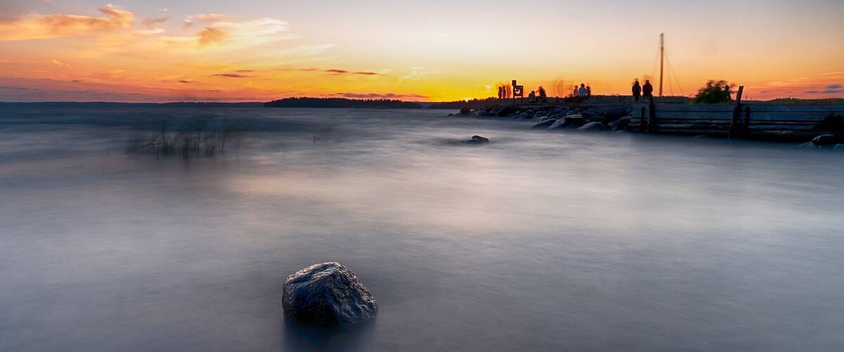 A sunset over a calm lake with a rocky outcrop in the foreground and a pier with people silhouetted against the sky.