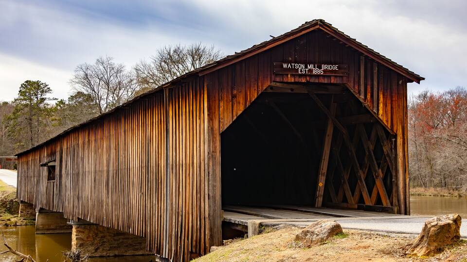 A wooden covered bridge with a sign reading "Watson Mill Bridge EST 1885" over a calm river.