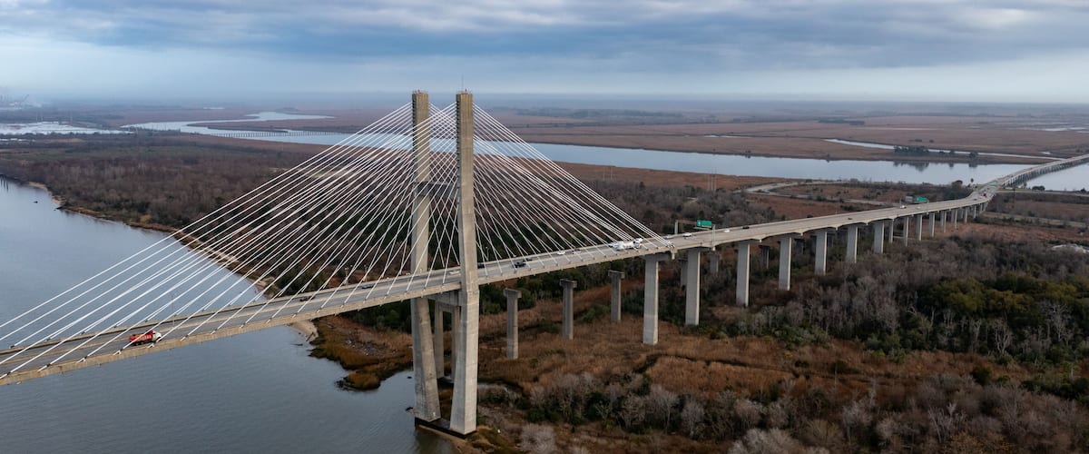 A large suspension bridge over a river, surrounded by fields and trees.