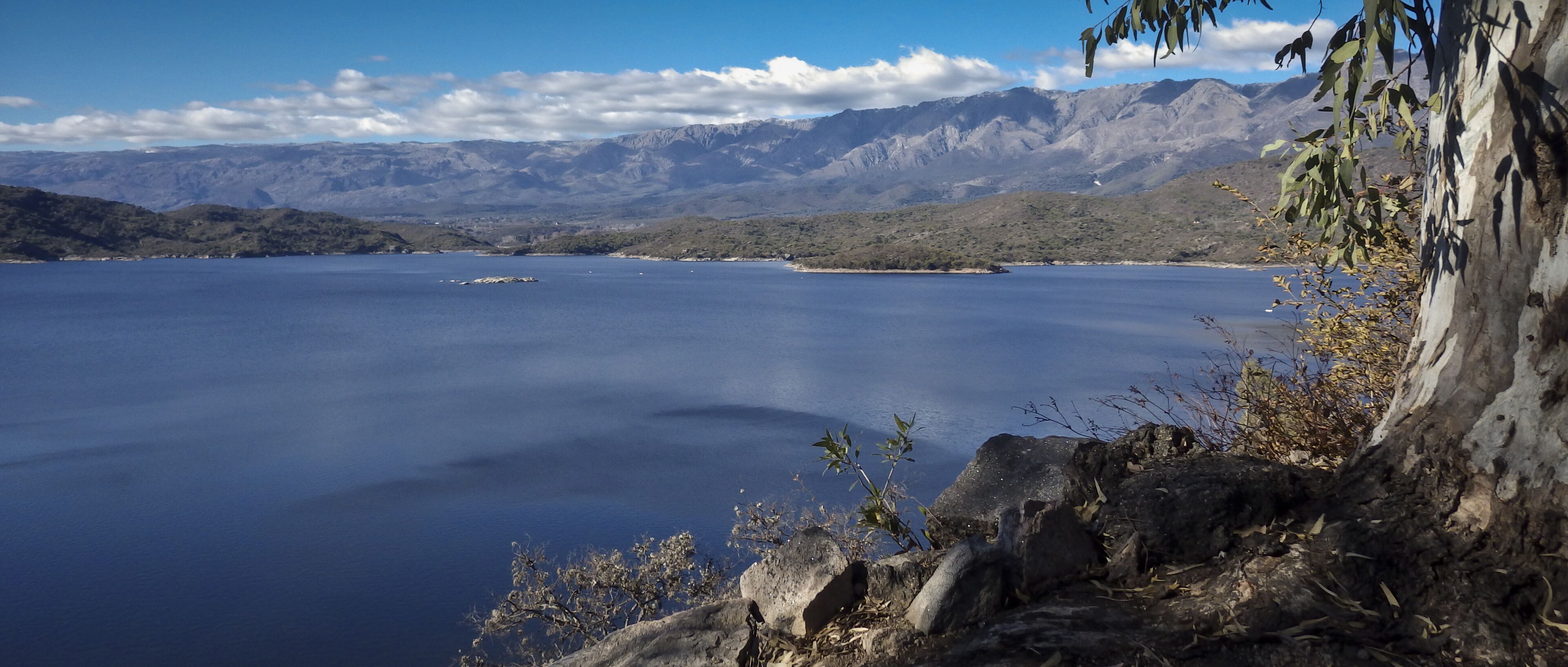A calm lake with mountains in the background and a rocky shoreline with sparse vegetation.