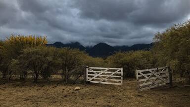 A landscape with a gate, dry bushes, and mountains under a cloudy sky.