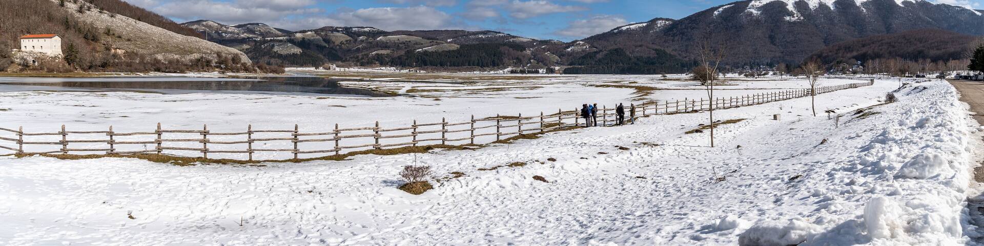 A snow-covered landscape with a wooden fence, a small building, and mountains in the background.