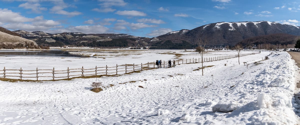 A snow-covered landscape with a wooden fence, a small building, and mountains in the background.