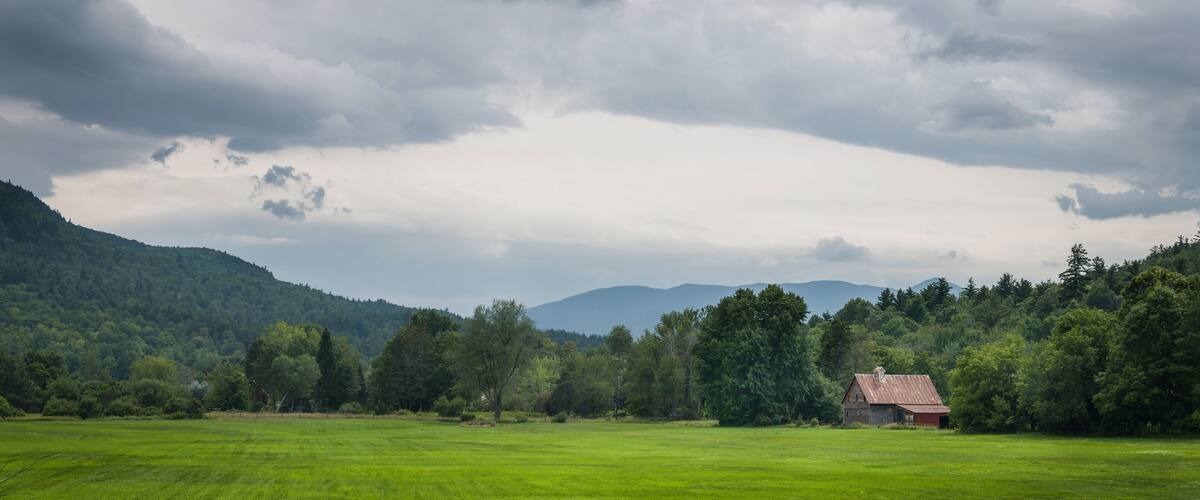 A green field with a small red-roofed building in the distance, surrounded by trees and hills under a cloudy sky.