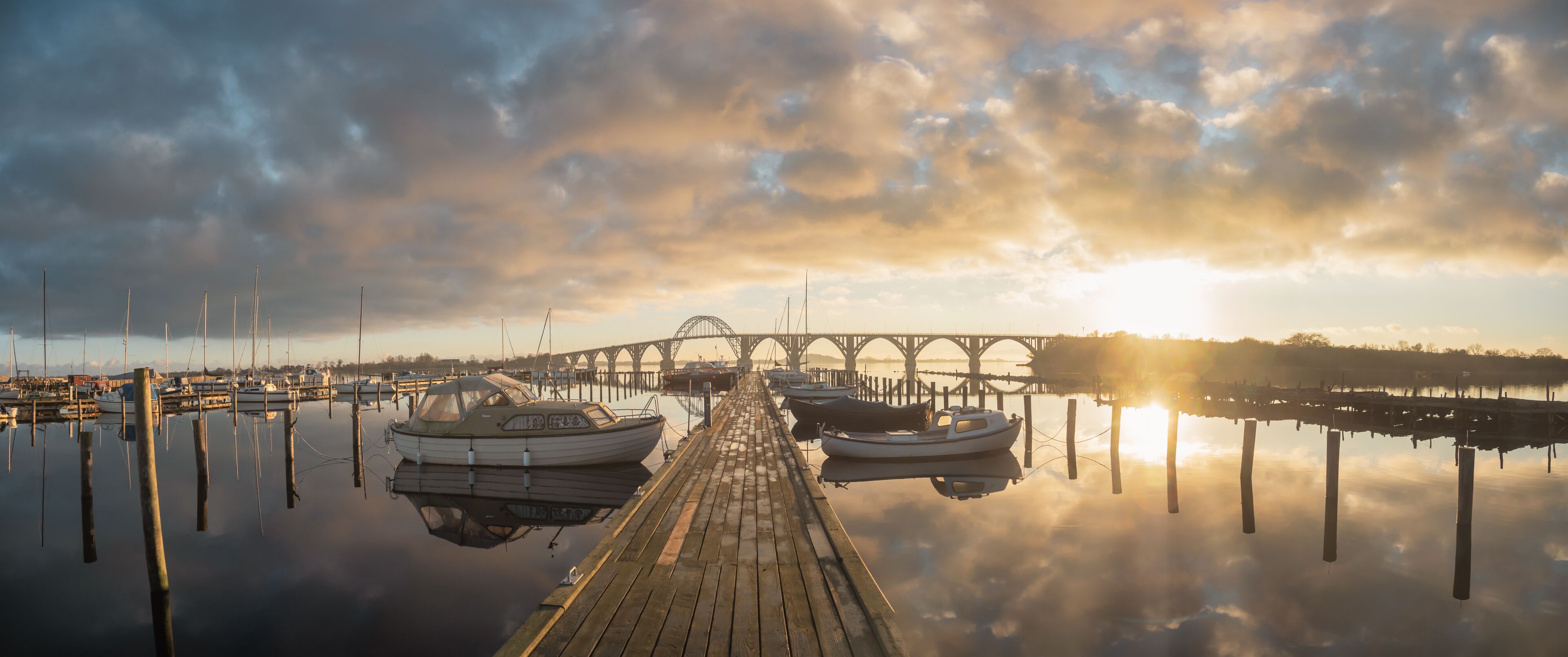 A wooden pier with boats docked, a bridge in the background, and a sunset over a calm body of water.