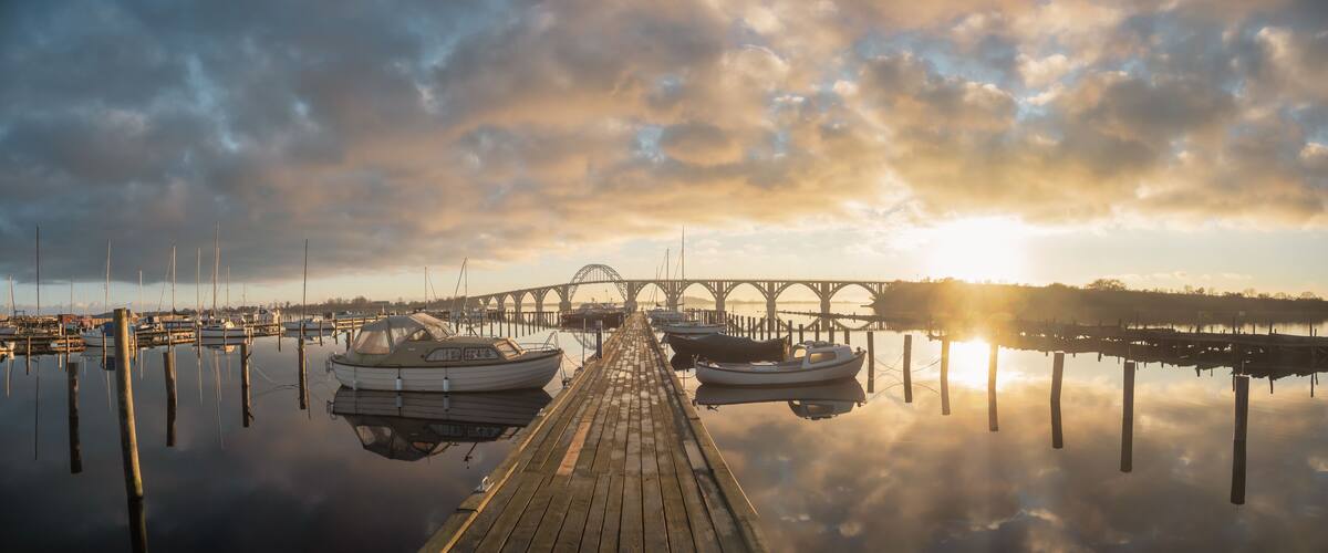 A wooden pier with boats docked, a bridge in the background, and a sunset over a calm body of water.