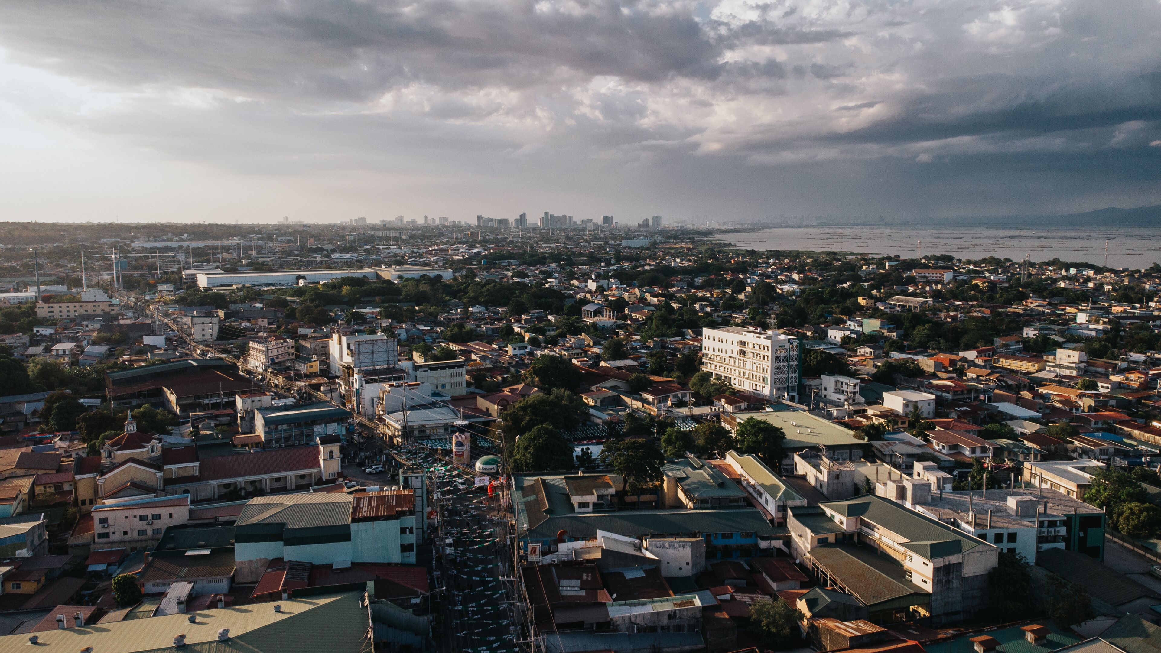 A coastal cityscape with dense urban development, a prominent waterfront, and a distant city skyline under a cloudy sky.