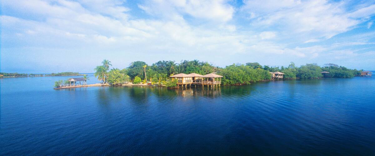 A tropical island with a clear blue sky, calm water, and a small house surrounded by lush greenery.