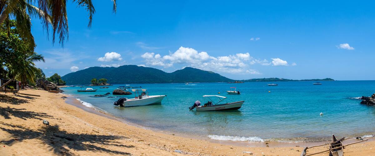 A beach with boats, clear blue water, and mountains in the background.