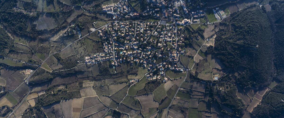 An aerial view of a rural area with a dense cluster of houses surrounded by fields and patches of forest.