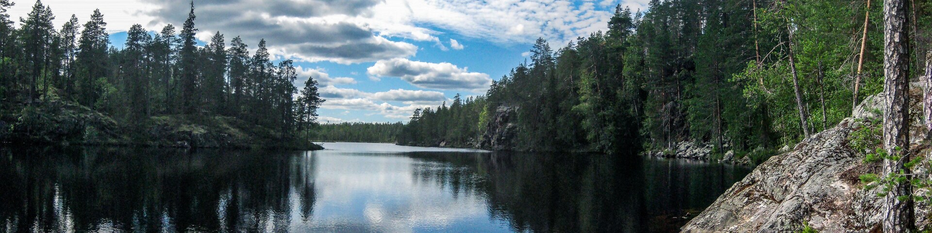 A calm lake surrounded by dense forest and a clear sky with scattered clouds.