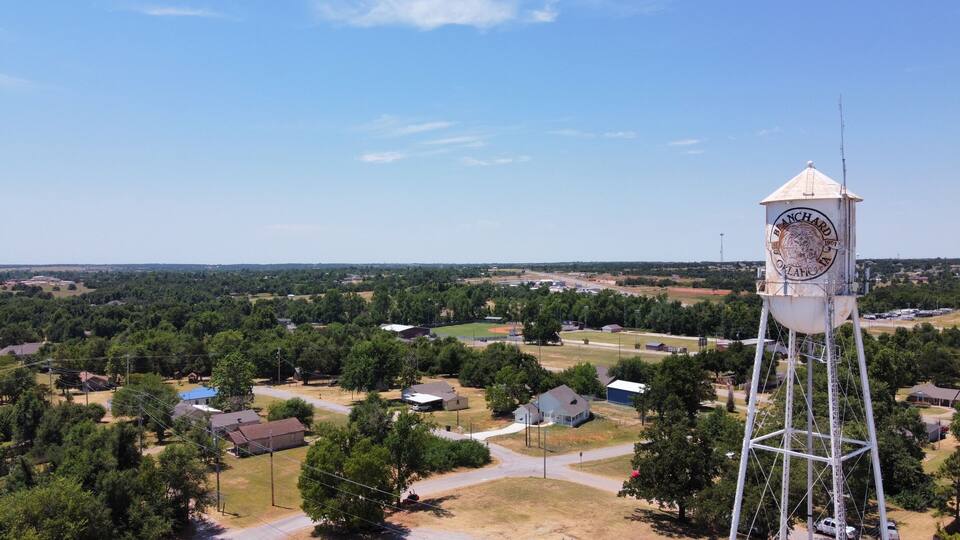 A water tower with a clock, surrounded by a rural landscape with houses and trees.