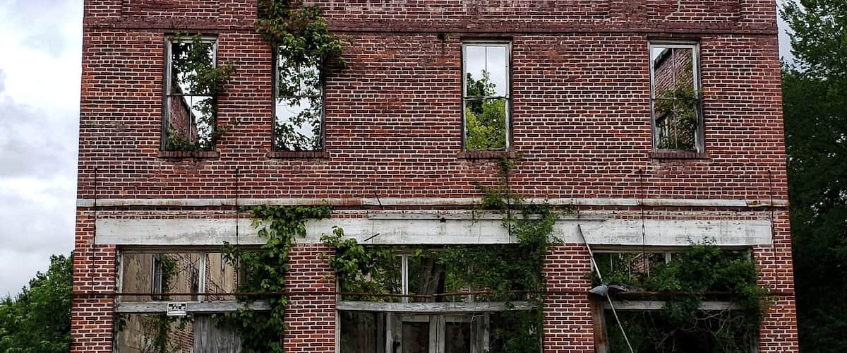 A large, abandoned brick building with boarded-up windows and overgrown vegetation.