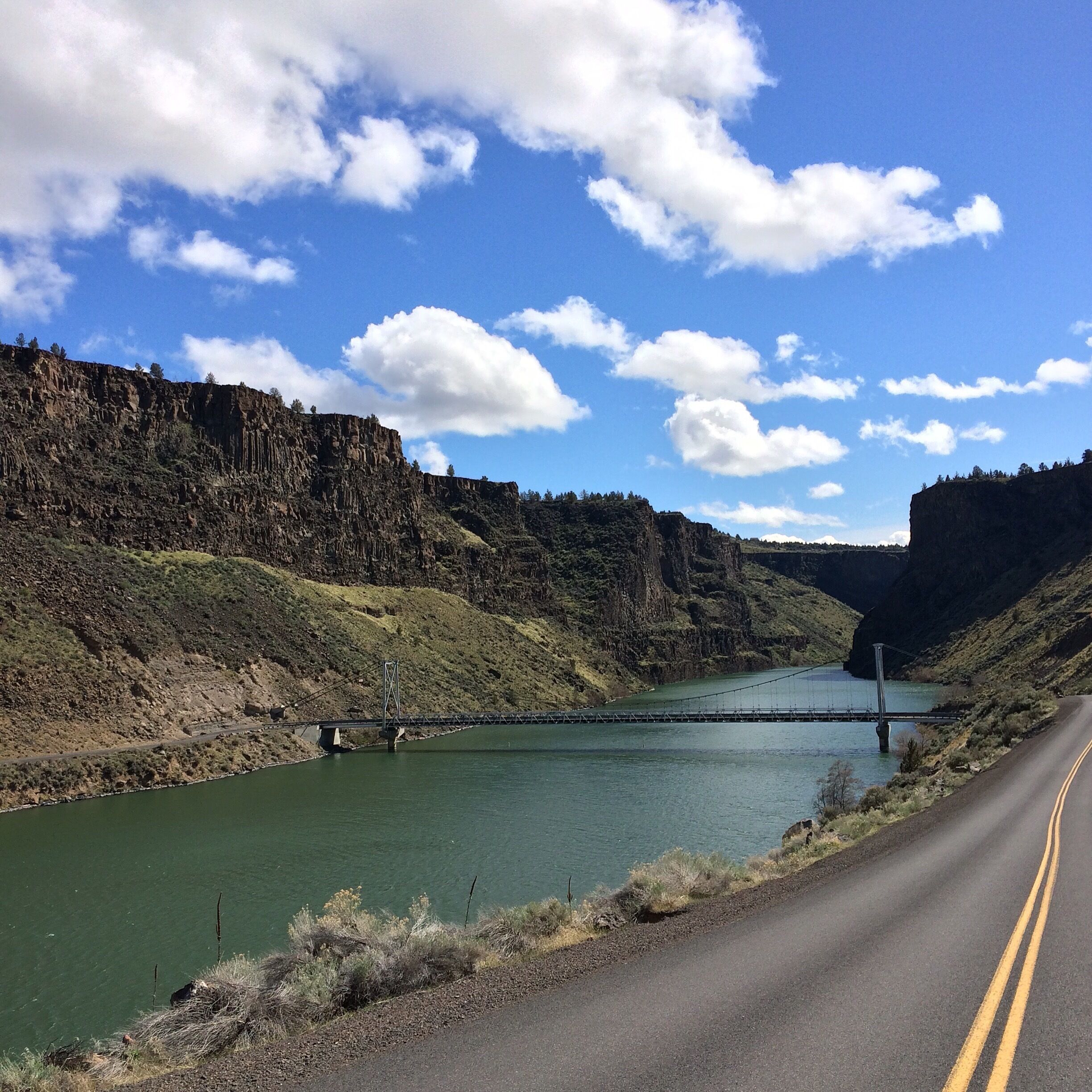 A river with a bridge, a road, and a mountainous landscape under a blue sky with clouds.