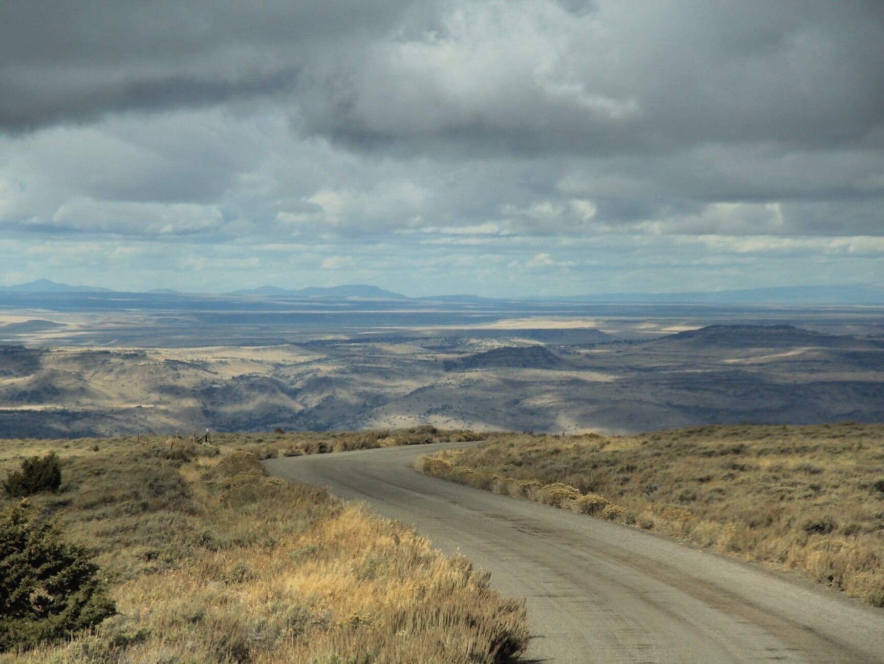 A winding dirt road through a dry, hilly landscape with distant mountains under a cloudy sky.