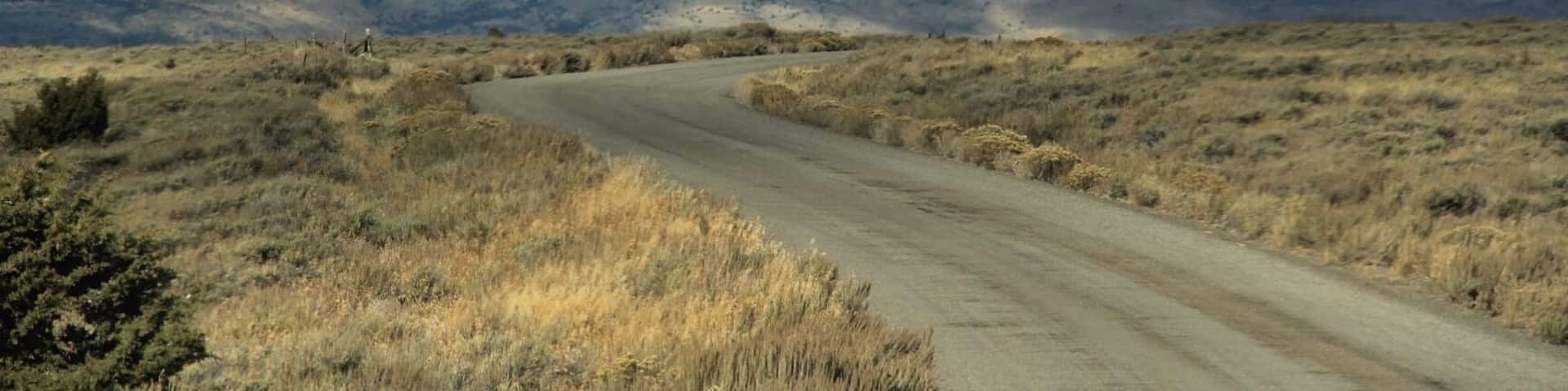 A winding dirt road through a dry, hilly landscape with distant mountains under a cloudy sky.