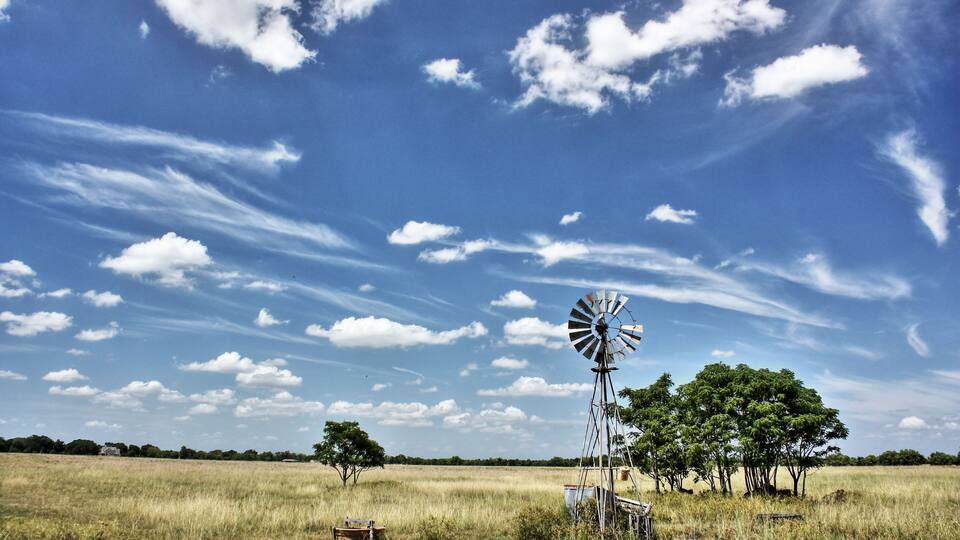 A windmill in a grassy field with trees and a clear blue sky.