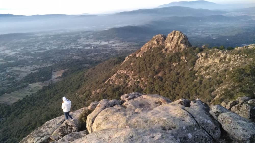 Une personne se tient sur un promontoire rocheux, d’où elle domine un paysage montagneux avec un sommet particulièrement imposant.