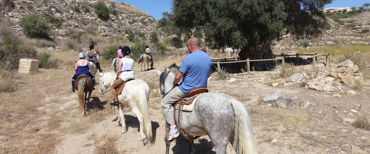 A group of people riding horses on a dirt path with rocky terrain and sparse vegetation.