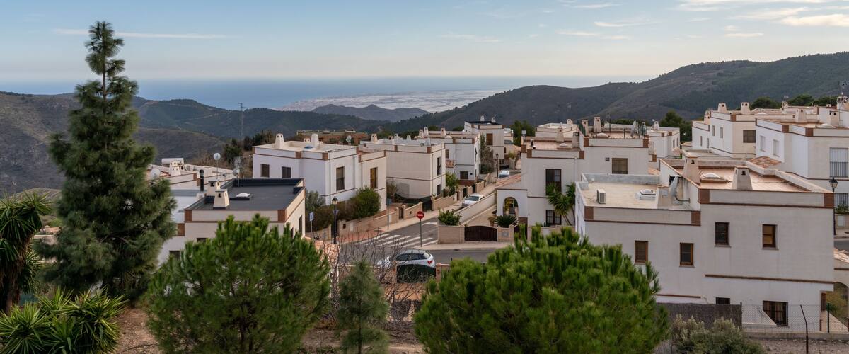 A coastal town with white buildings, green trees, and a view of the sea and distant hills.