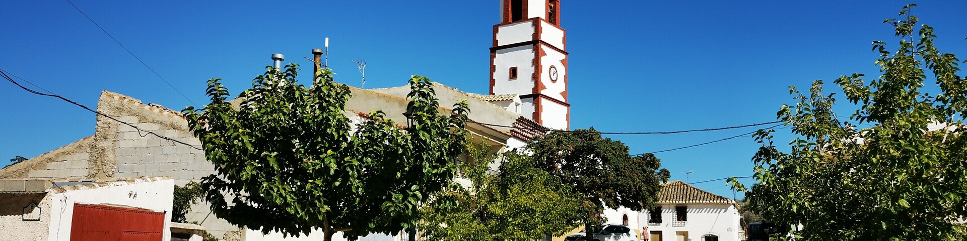 A church with a clock tower, benches, and trees in a sunny courtyard.