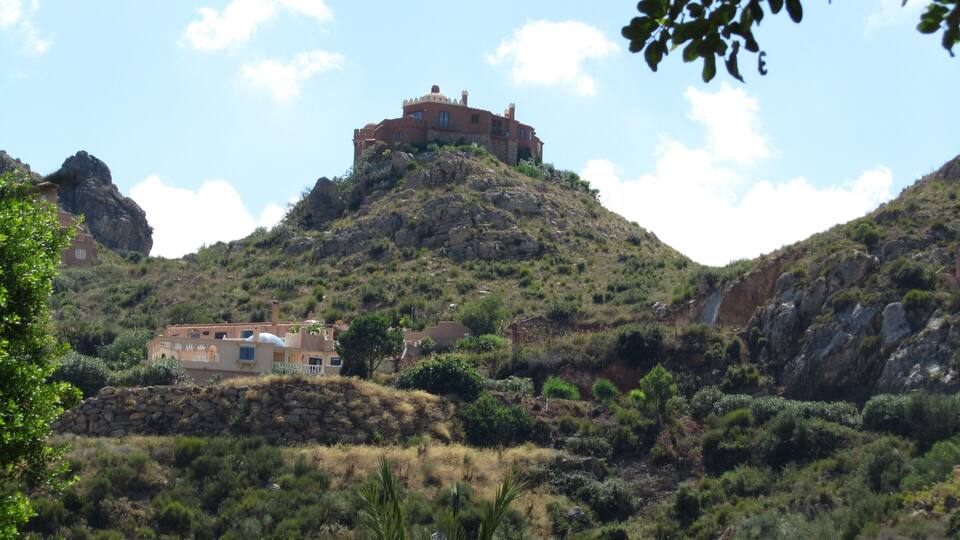 A hilltop house with a red roof, surrounded by greenery and rocky terrain.