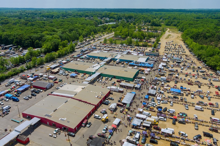An aerial view of a large parking lot with numerous vehicles and temporary structures, surrounded by dense green forest.