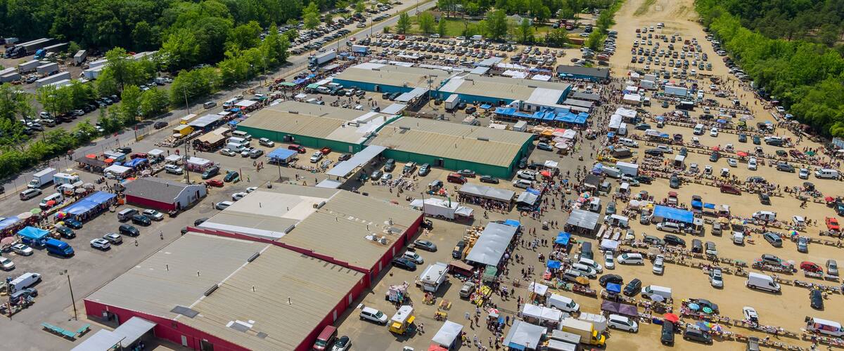 An aerial view of a large parking lot with numerous vehicles and temporary structures, surrounded by dense green forest.