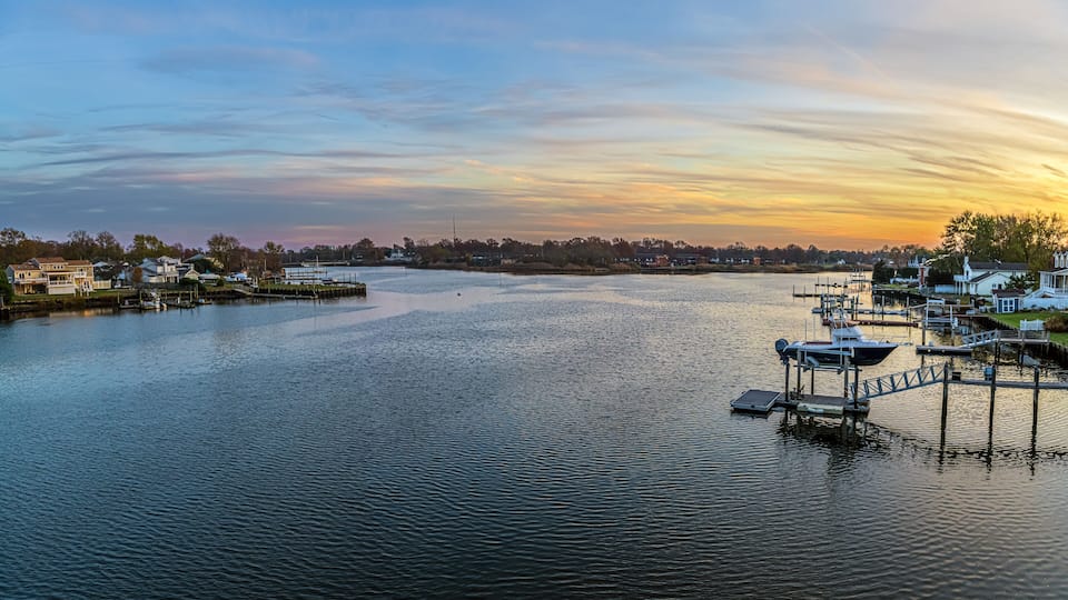 A marina with boats docked, a calm river, and houses along the shore.