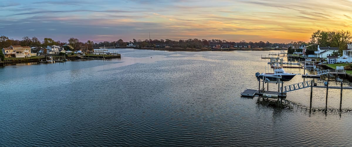 A marina with boats docked, a calm river, and houses along the shore.