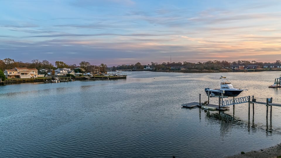 A calm river with boats docked at a pier, surrounded by houses and trees.