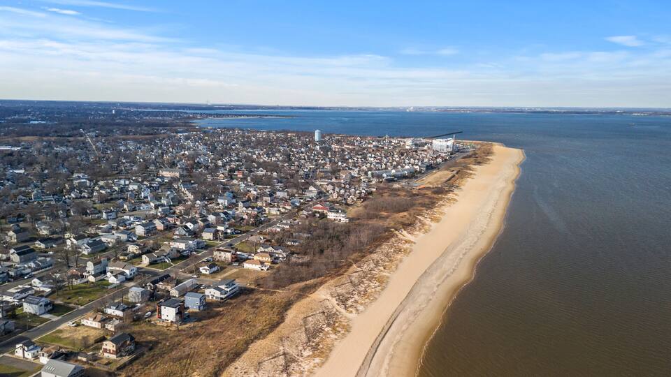 An aerial view of a coastal town with a sandy beach, dense residential area, and a large body of water.