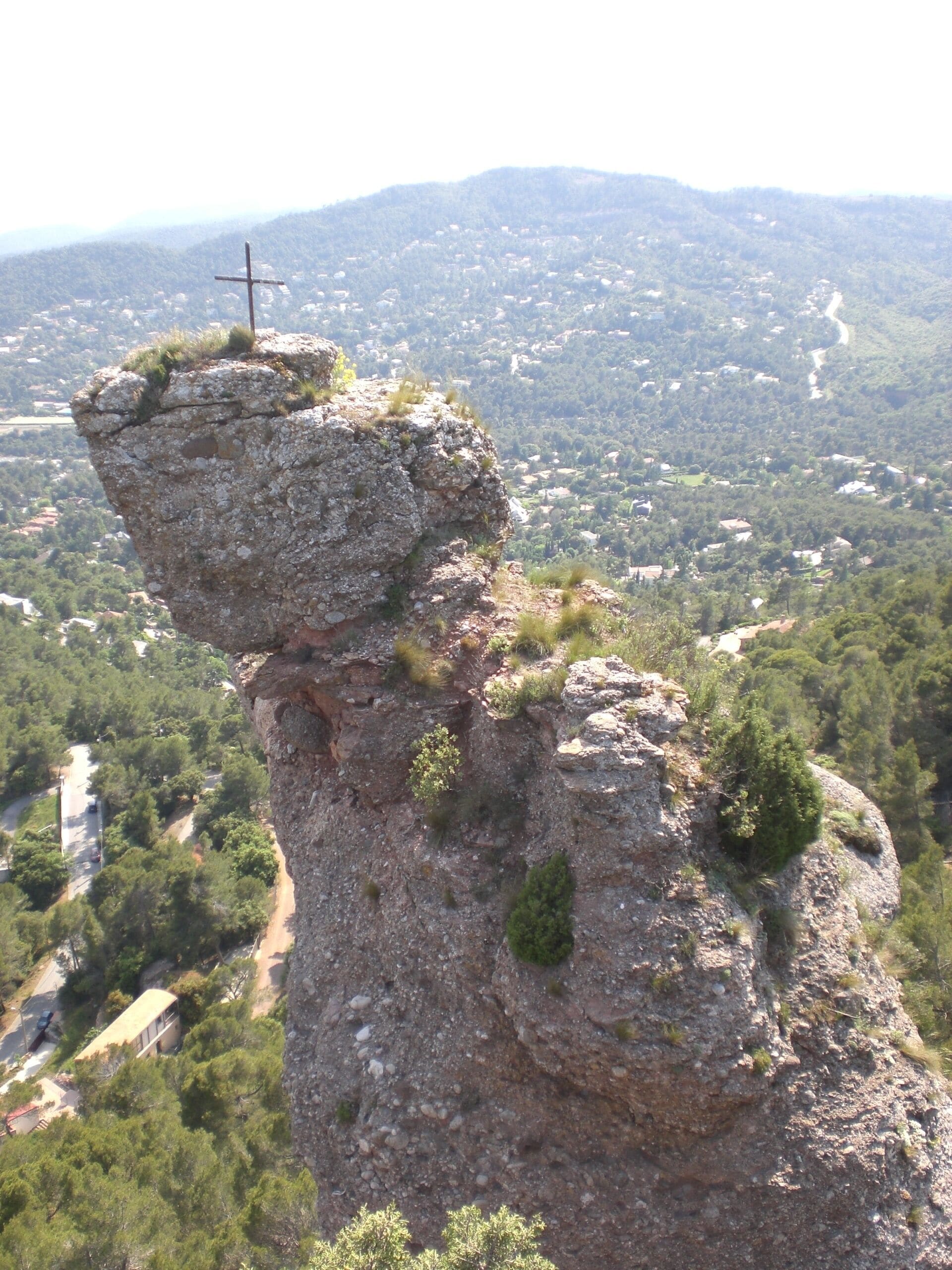 A rock formation with a cross on top, surrounded by greenery and a distant village.