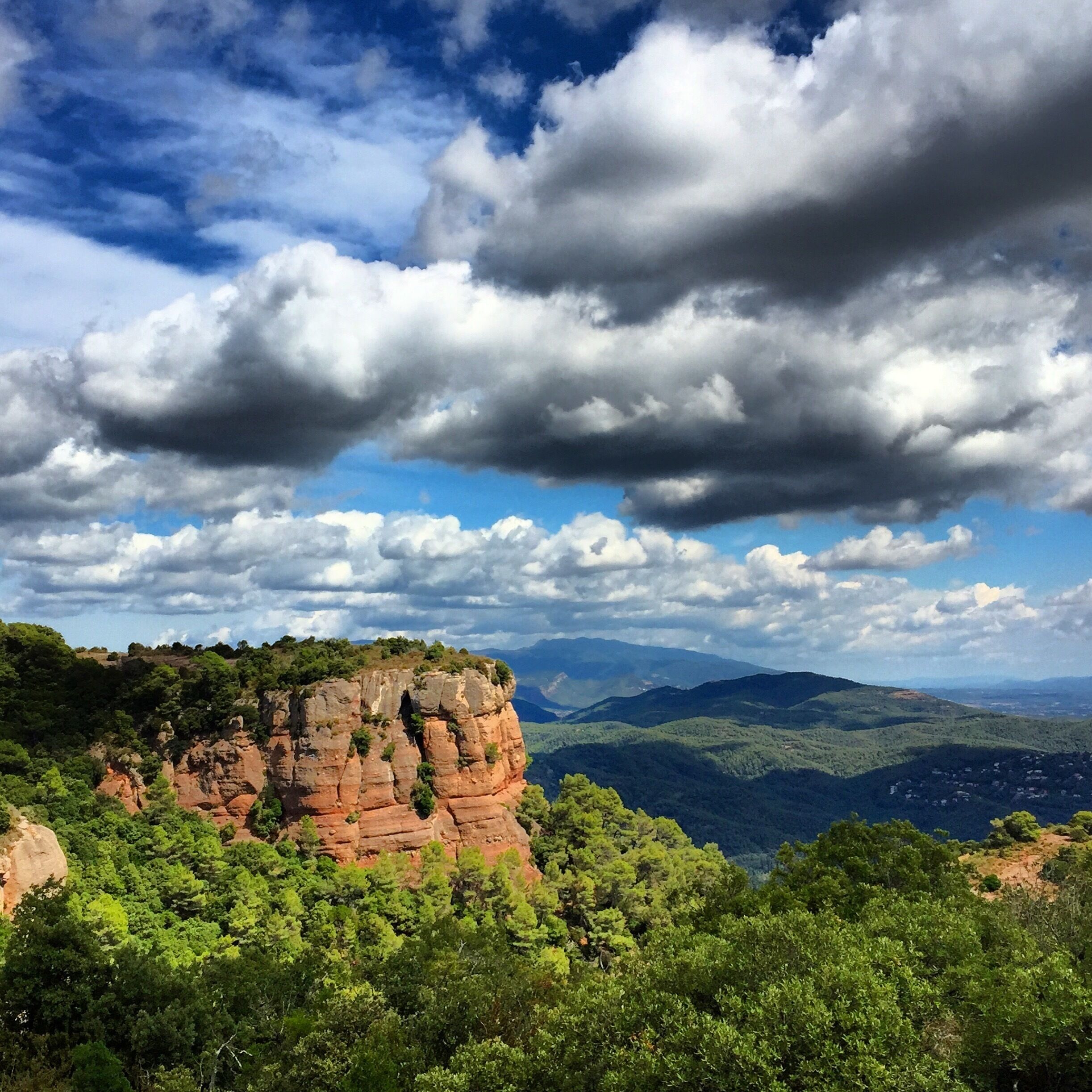 A rocky cliff with green vegetation, a cloudy sky, and distant mountains.