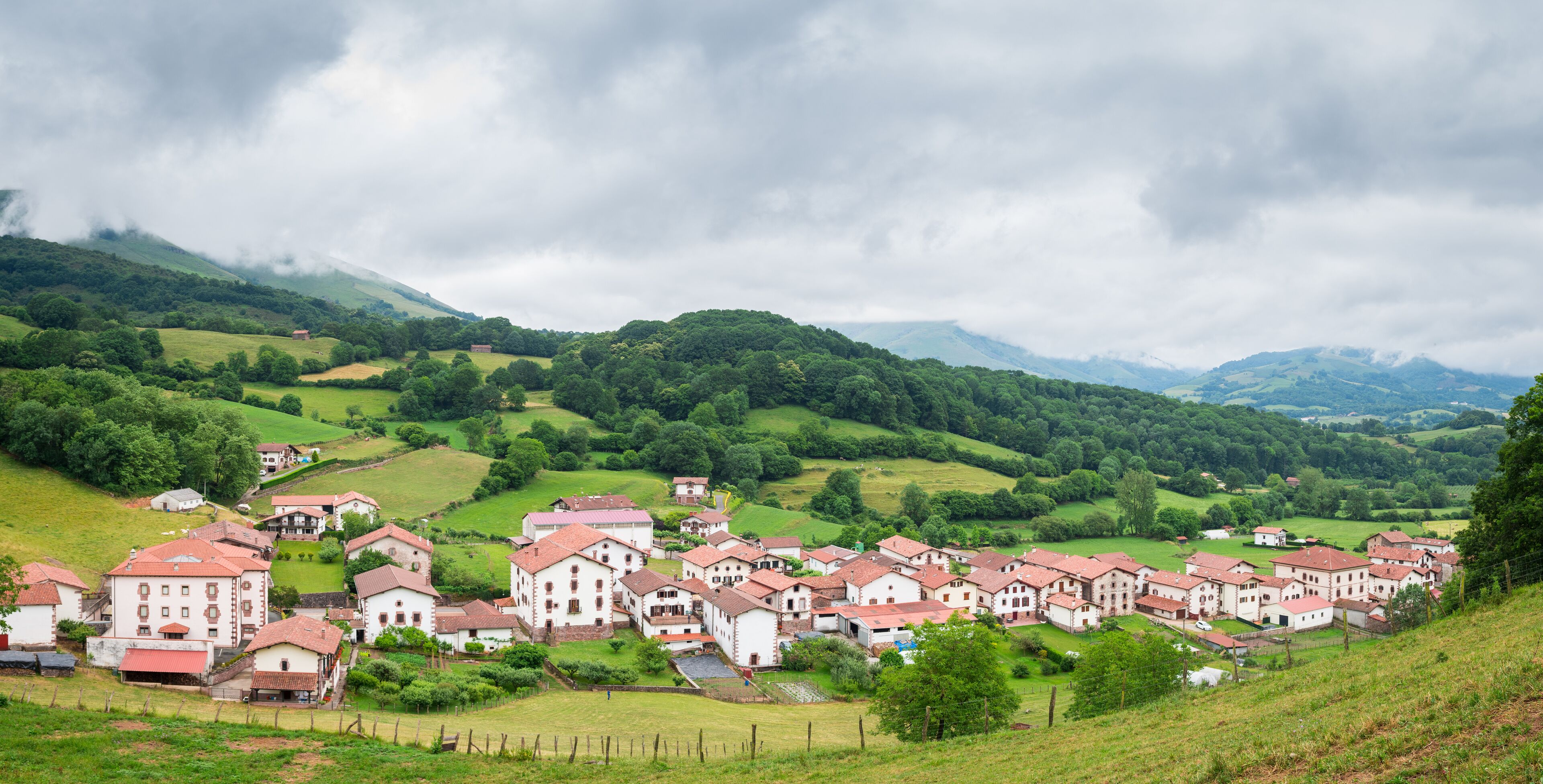A village with red-roofed buildings nestled in green hills under cloudy skies.