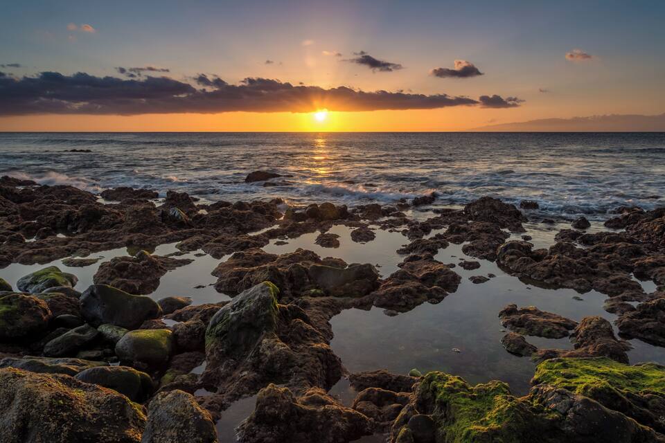A rocky shoreline at sunset with the sun setting over the ocean.