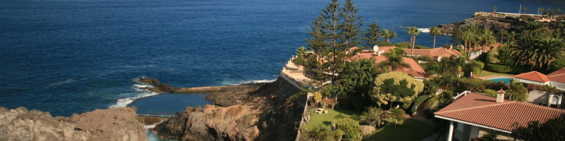 A coastal landscape with rocky cliffs, a clear blue sea, and a residential area with houses and greenery.