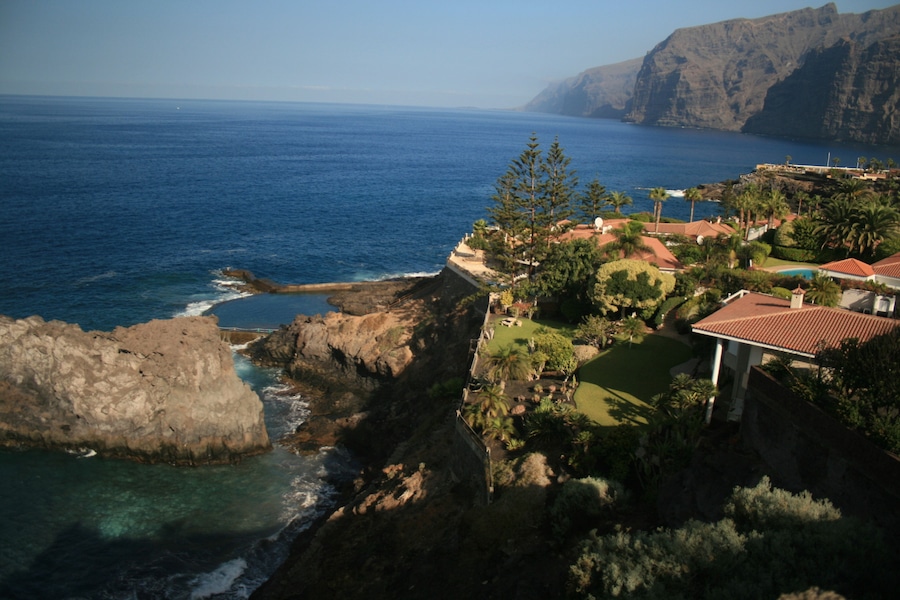 Un paisaje costero con acantilados rocosos, mar azul cristalino y una zona residencial con casas y vegetación.