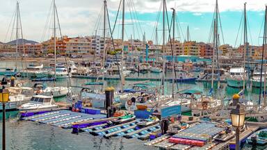A marina with numerous boats docked, a beach, and buildings in the background.
