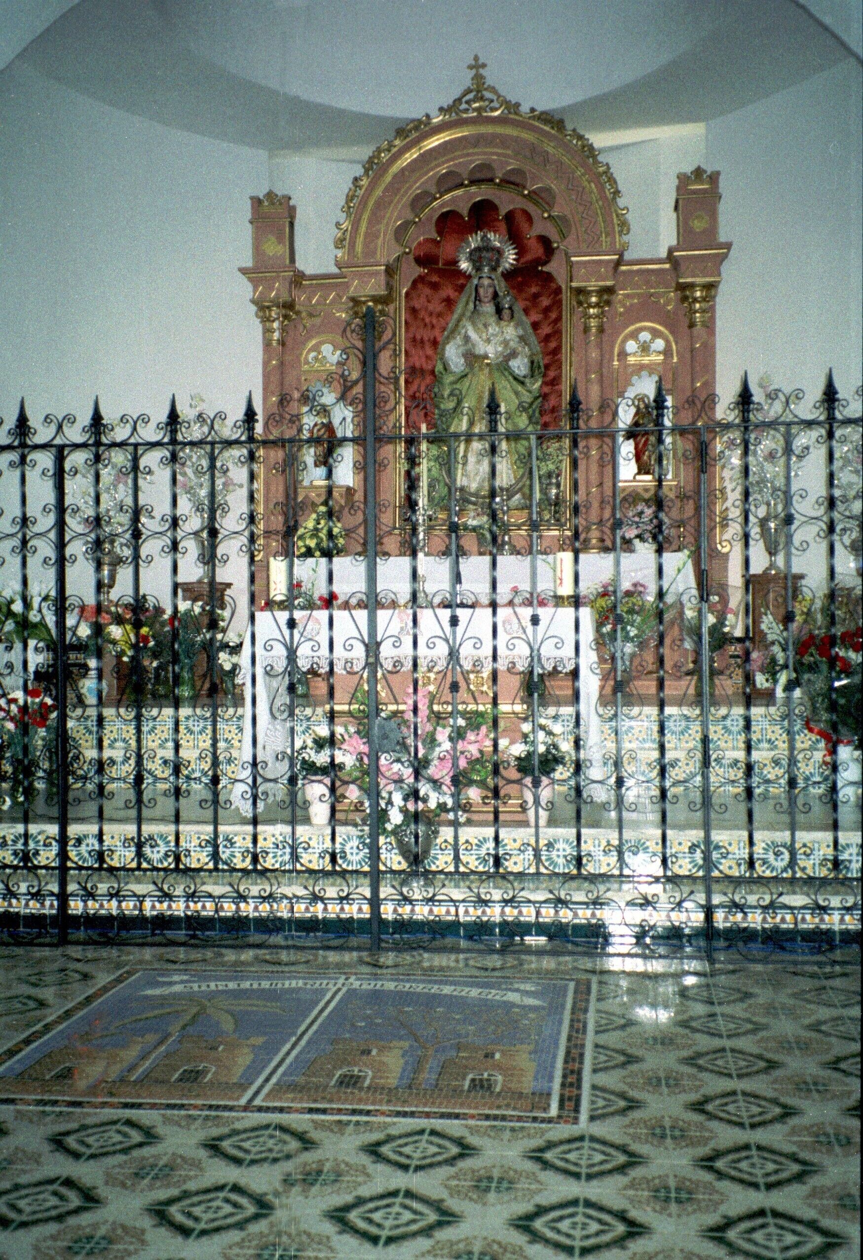A religious statue behind a wrought iron fence, with patterned floor tiles and floral arrangements.