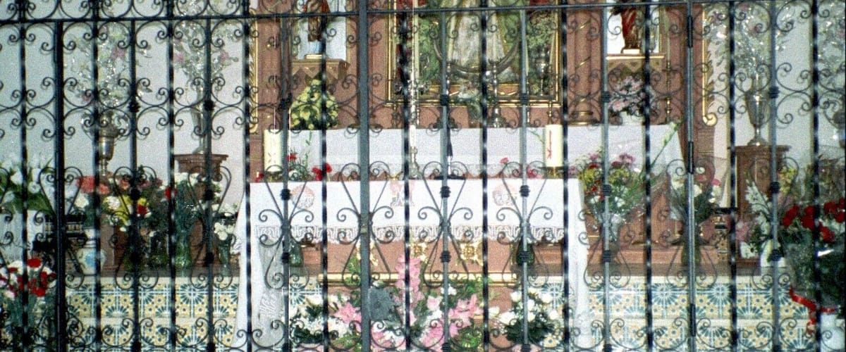 A religious statue behind a wrought iron fence, with patterned floor tiles and floral arrangements.