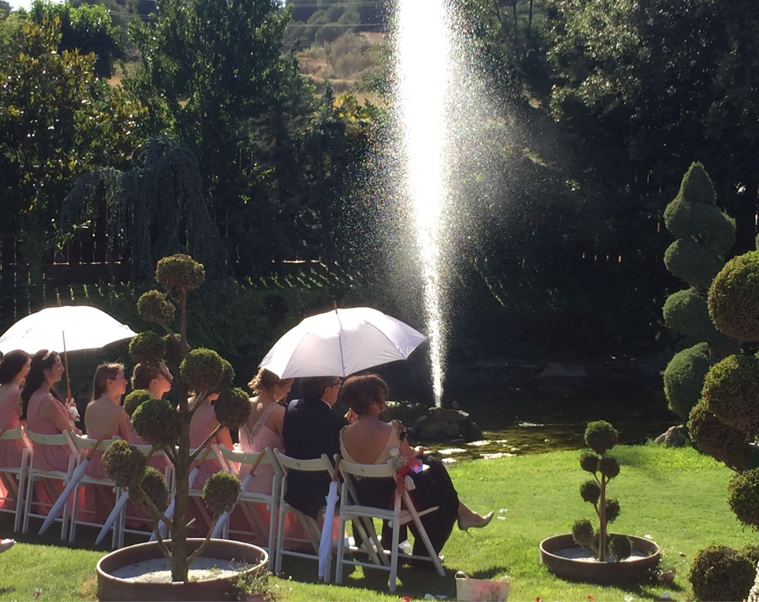 A group of people seated on white chairs with umbrellas, facing a fountain in a garden.