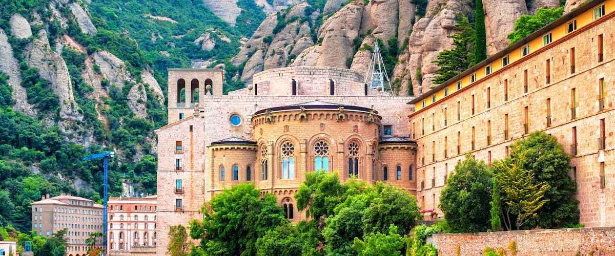 A historic building with a circular central section and a surrounding rectangular structure, set against a backdrop of rugged, rocky mountains and lush greenery.